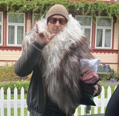 a man in a furry coat and hat making the peace sign with his hand while standing next to a white picket fence