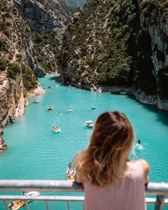 a woman standing on a balcony looking at boats in the blue water and mountains behind her