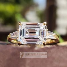 an emerald cut diamond ring with three baguets on the band, set in yellow gold