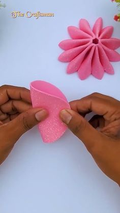 two hands holding a pink paper flower on top of a white table next to flowers