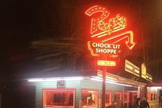 people are standing in front of a shop at night with the neon sign lit up