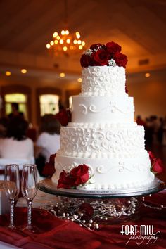 a white wedding cake with red roses on top and wine glasses in the foreground