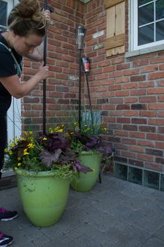 a woman is watering plants in front of a brick building with two large green planters