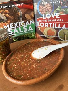 a bowl of salsa next to two cookbooks and a spoon on a table top