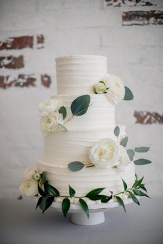 a wedding cake with white flowers and greenery on top is displayed in front of a brick wall