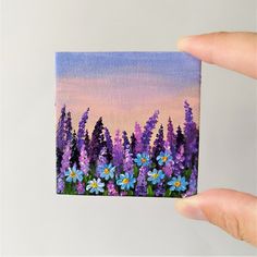 a hand holding up a small painting of purple and blue flowers in front of a pink sky