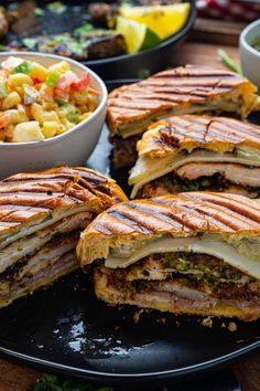 several sandwiches cut in half on a black plate with bowls of fruit and salad behind them