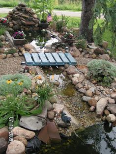 a small pond with rocks and water features an american flag in the background, surrounded by flowers