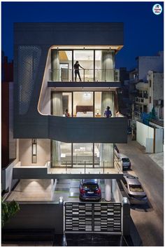 an architecturally designed house at night with people standing on the balcony and cars parked outside