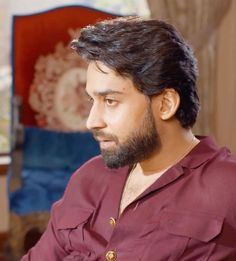 a man with a beard sitting in a chair looking off to the side and wearing a red shirt