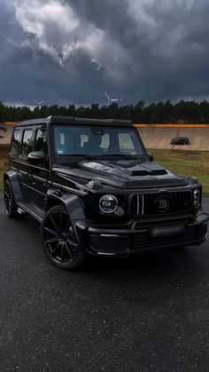 a black mercedes g class is parked on the side of the road in front of dark clouds