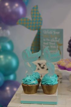 two cupcakes with blue frosting are sitting on a table in front of balloons