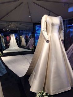 a white wedding dress on display in a glass case with flowers and other gowns behind it