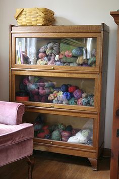 a pink chair sitting in front of a wooden cabinet filled with balls of knitting yarn