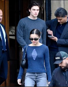 a woman in a blue sweater and jeans is looking at her cell phone while other people look on