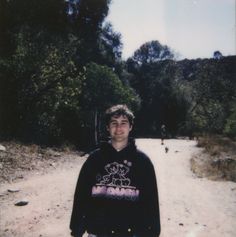 a young man standing in the middle of a dirt road with trees and bushes behind him