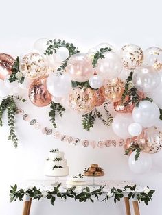 a table topped with lots of balloons and greenery next to a wall covered in white and pink balloons