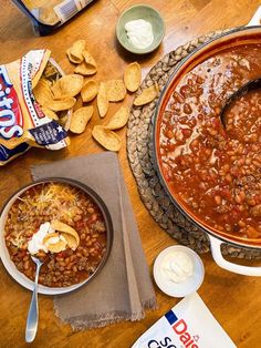 a table topped with bowls of chili and cornbreads next to chips on top of a wooden table