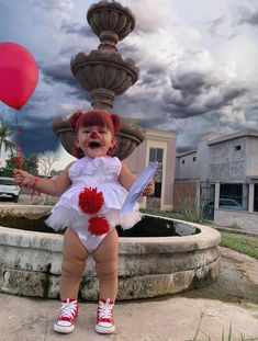 a toddler in a costume standing next to a fountain and holding a red balloon