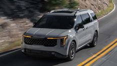 a silver suv driving down the road in front of a mountain side with trees and bushes