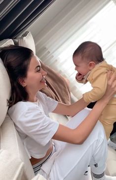 a woman holding a baby while sitting on top of a white couch next to a window