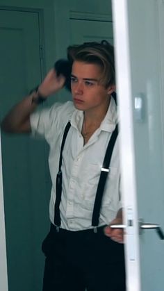 a young man in suspenders and a white shirt is looking at himself through the mirror