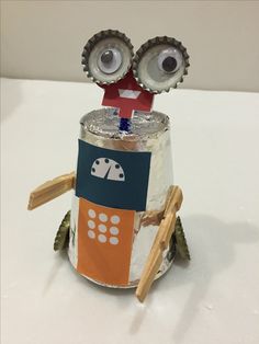 a paper mache robot with two eyes sitting on top of a white table next to a pair of wooden spoons