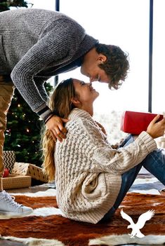 a man and woman kissing while sitting on the floor in front of a christmas tree