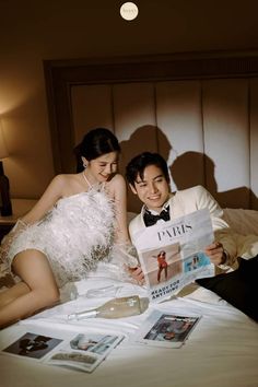 a man and woman sitting on top of a bed next to each other reading a magazine