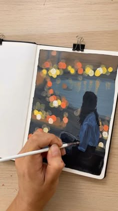 a person is holding a paintbrush and drawing on an open book with blurry lights in the background