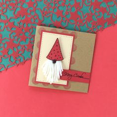 a handmade card with a red hat and tassel on the front, sitting on a pink background