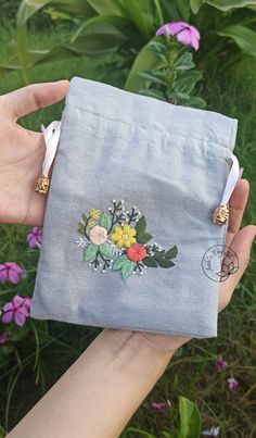 a hand holding a small blue pouch with flowers on the front and side, in front of some purple flowers