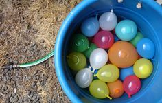 a bucket filled with lots of colorful balls on top of a dirt ground next to a hose