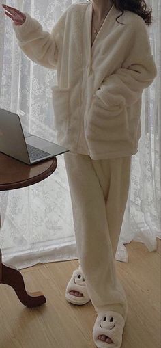 a woman in white pajamas standing next to a table with a laptop computer on it