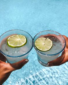 two people holding up glasses with lemons and limes in them next to a swimming pool