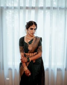 a woman in a green and gold sari standing next to a window with sheer curtains