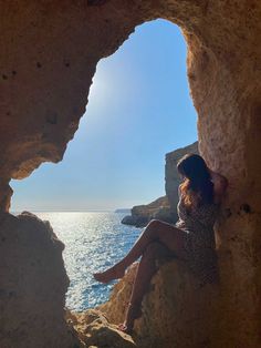 a woman sitting on top of a rocky cliff next to the ocean under a blue sky