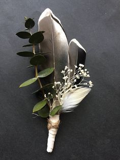 a feather and flower boutonniere on a black surface with white flowers, greenery and leaves