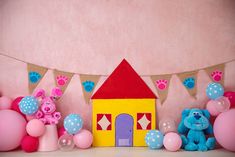 a blue teddy bear sitting in front of a toy house surrounded by pink and blue balloons