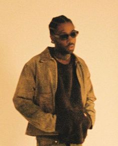 a man standing in front of a white wall with his hands in his pockets and wearing sunglasses