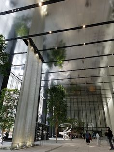 people are walking around in front of a building with glass walls and trees on both sides