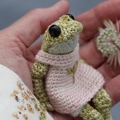 a hand holding a small crocheted frog in a sweater and pants with two tiny flowers