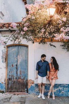 a man and woman standing in front of a building with pink flowers on the wall