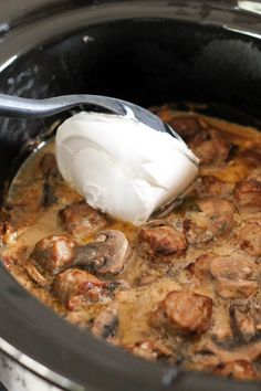 a spoon is in a slow cooker filled with meatballs and onions, topped with sour cream