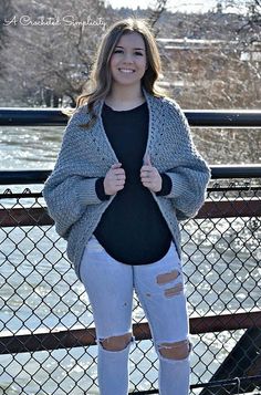 a woman standing in front of a fence wearing ripped jeans and a cardigan sweater