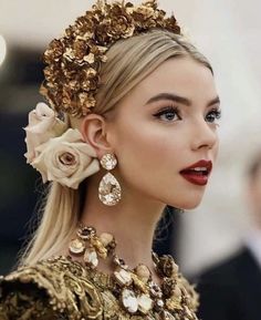 a woman with long blonde hair wearing a gold dress and flower in her hair is looking off to the side