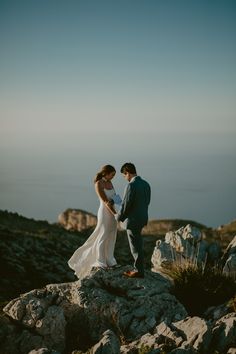 a bride and groom standing on top of a rocky hill holding each other's hands