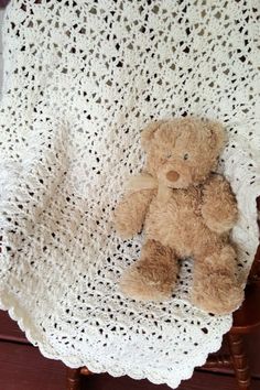 a brown teddy bear sitting on top of a white crocheted blanket next to a wooden chair