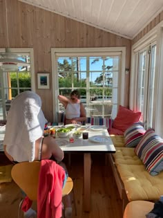 two women are preparing food in a small room with wooden walls and flooring, while another woman stands at the window