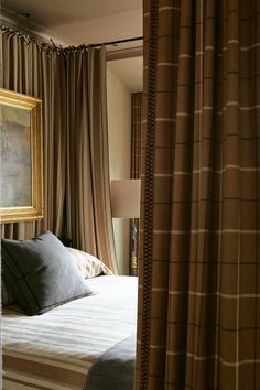 a bed sitting next to a window with curtains on the sides and a painting hanging above it